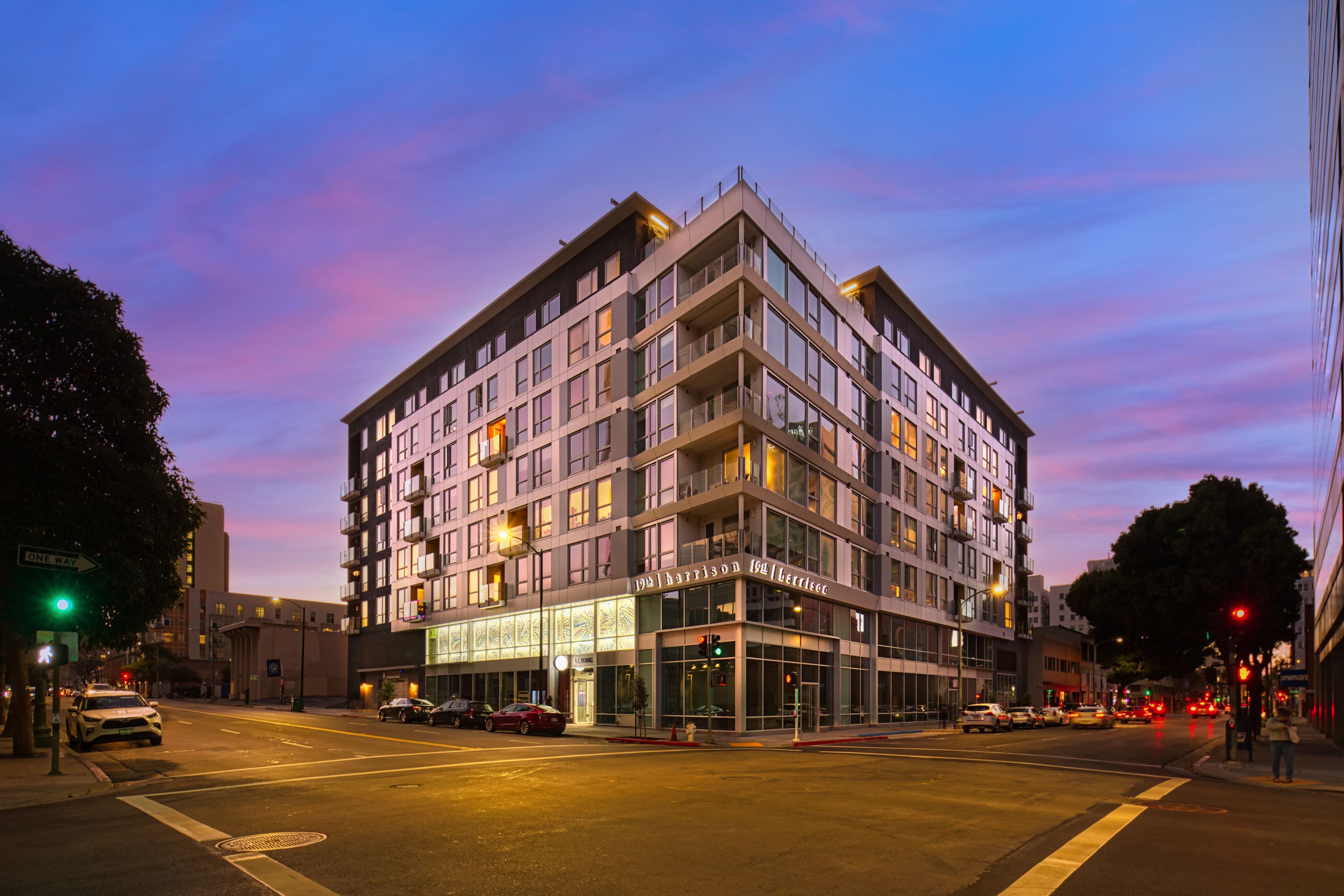 19th & Harrison at dusk, a seven-story apartment building with a glass corner at a downtown Oakland intersection