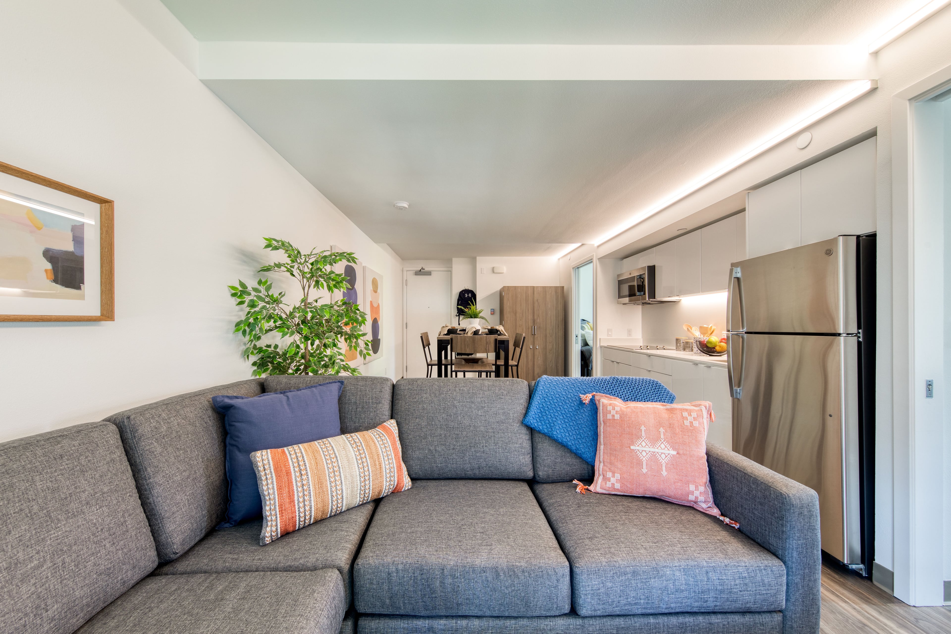 Living room with a grey sectional and a kitchen with stainless refrigerator beyond