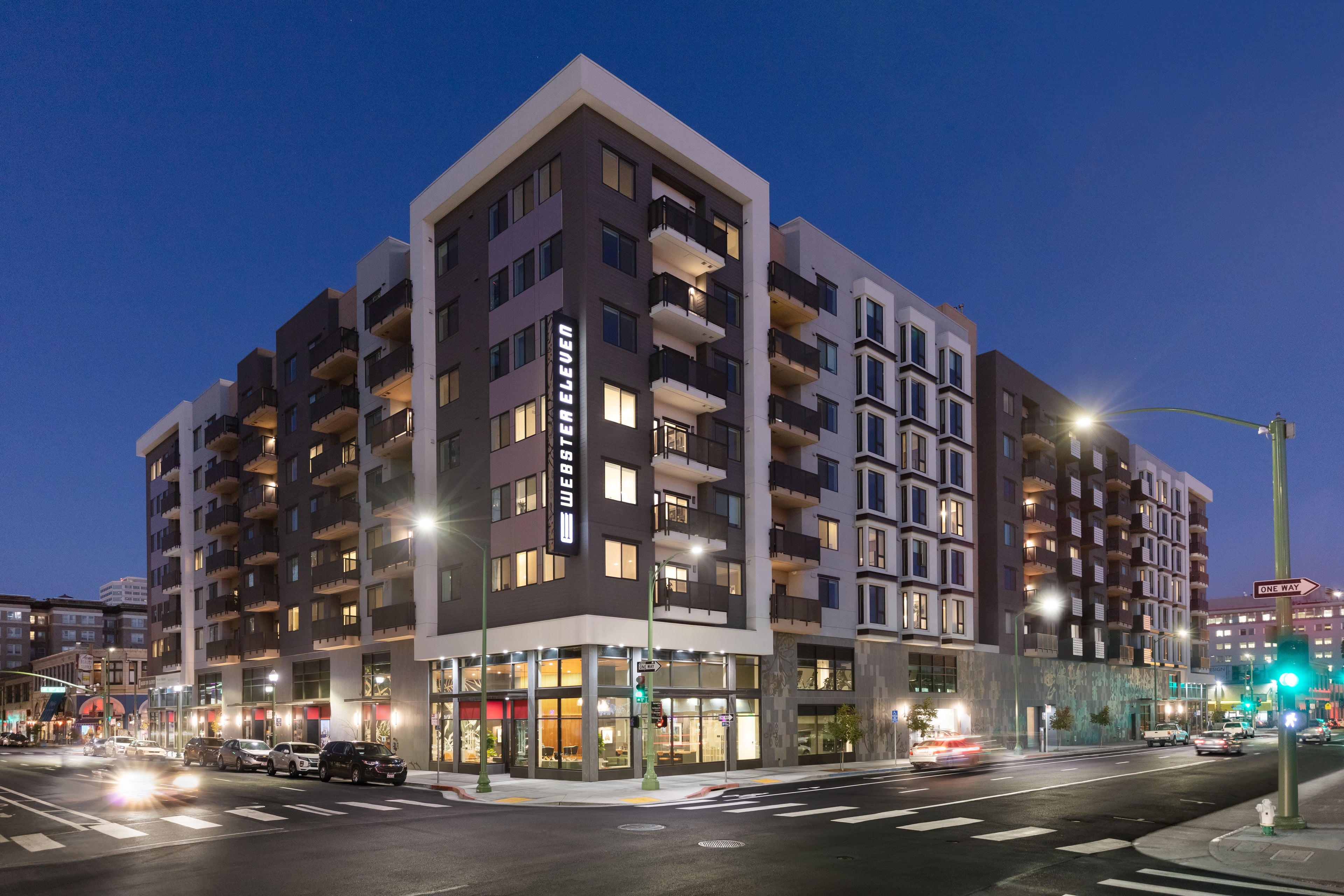 Webster Eleven at dusk, an eight-story apartment building on a downtown Oakland corner with lit ground-floor retail