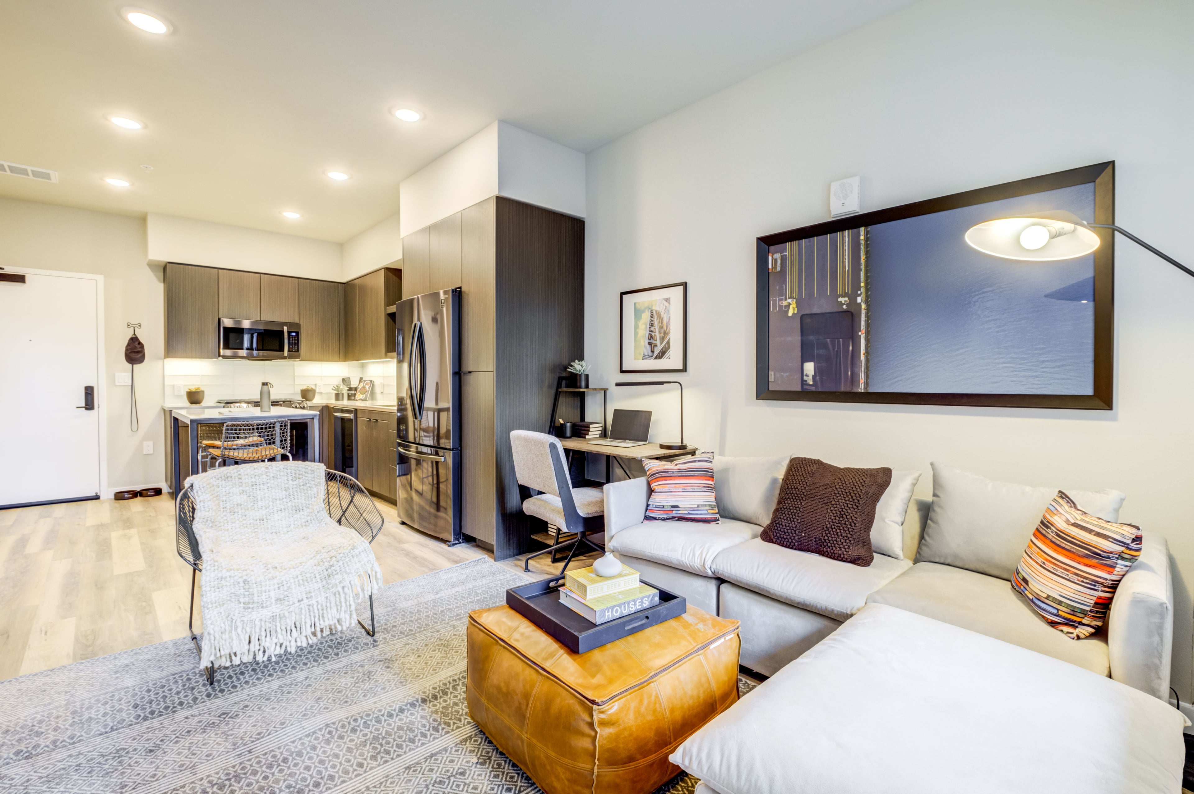 Living room with a white sectional, leather ottoman, and kitchen with dark cabinets beyond