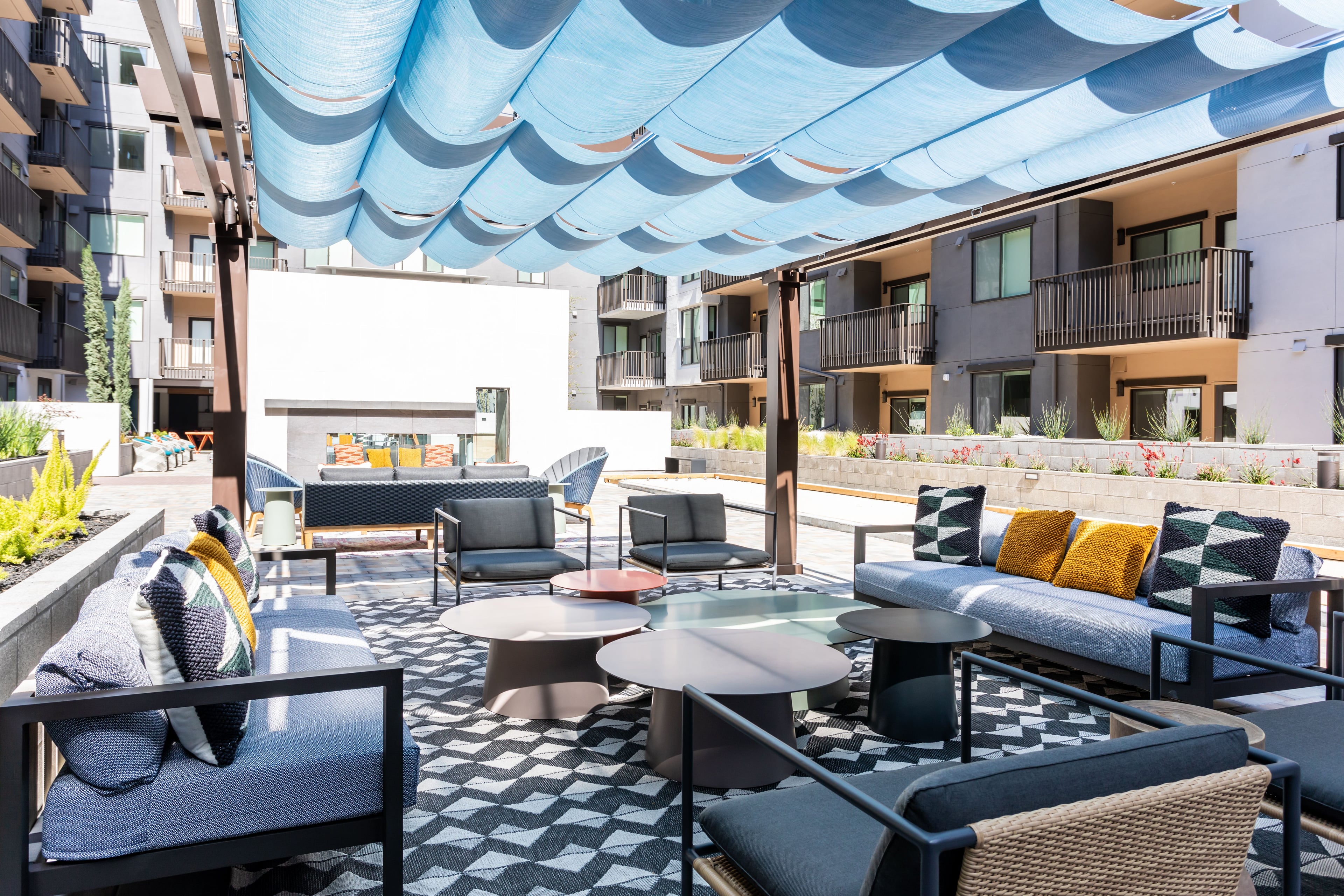 Covered courtyard lounge with sectional seating, patterned rugs, and a blue shade sail