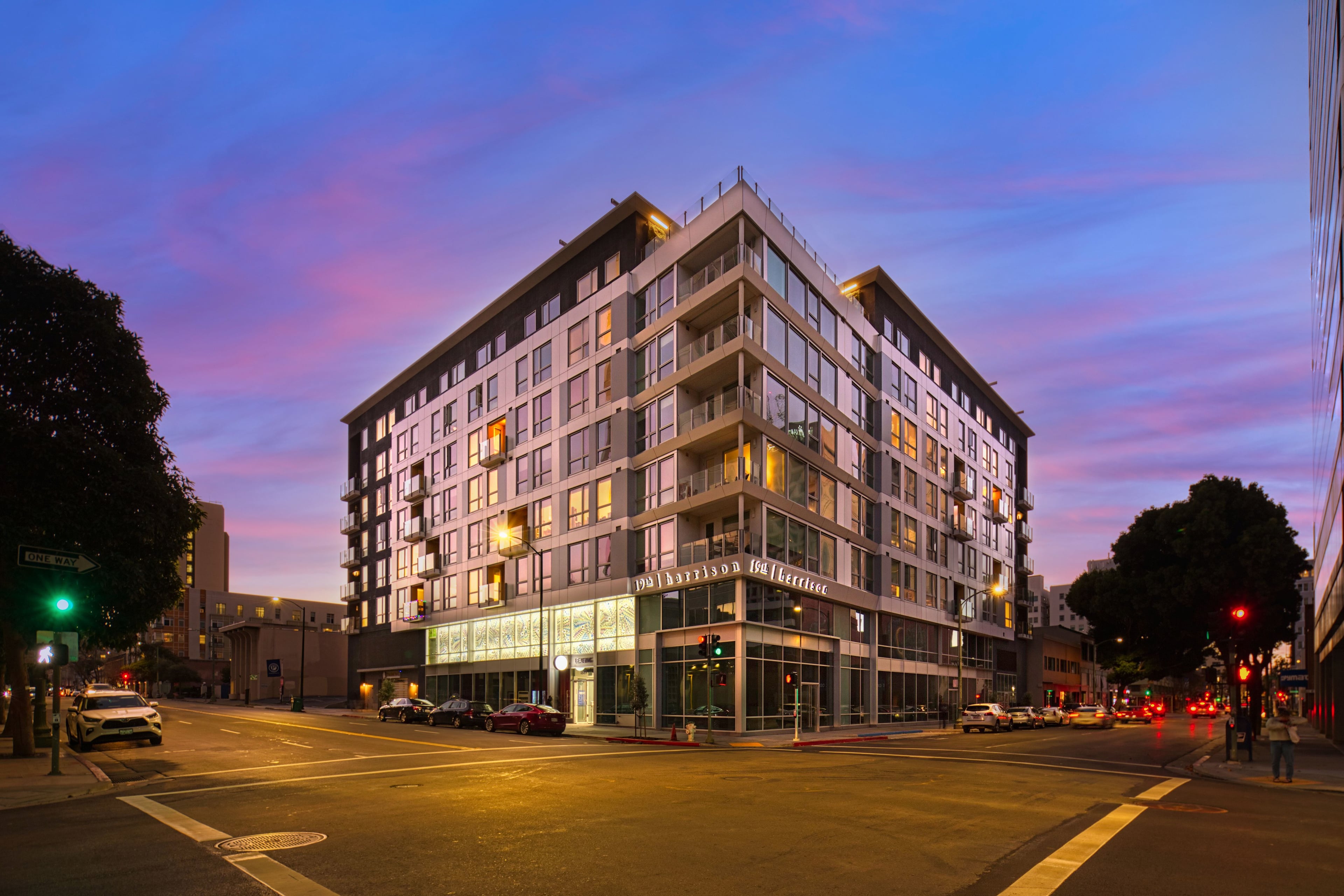 19th & Harrison at dusk, a seven-story apartment building with a glass corner at a downtown Oakland intersection