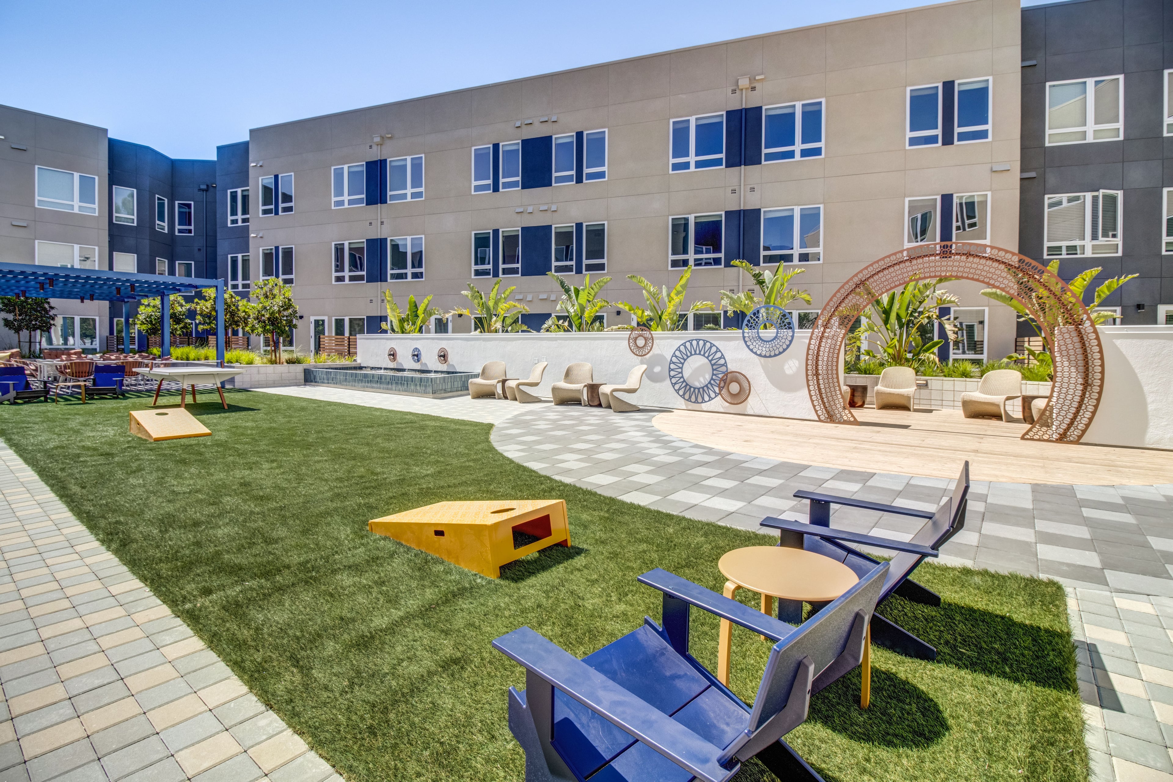 Courtyard with turf, cornhole boards, blue Adirondack chairs, and a metal arch sculpture
